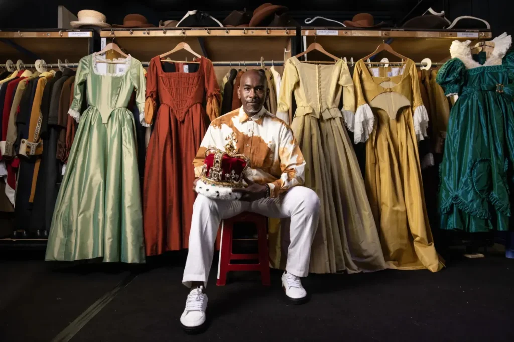 A man in white clothes sits on a red stool holding a crown, surrounded by historical dresses hanging on racks behind him, with hats and costumes arranged on the shelves above.