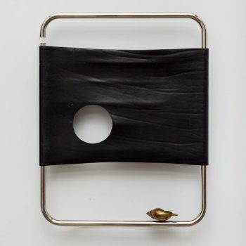 A minimalist object with a rectangular metal frame, a stretched black band featuring a circular hole, and a small brass bird-shaped figure attached to the bottom right corner. The background is white.