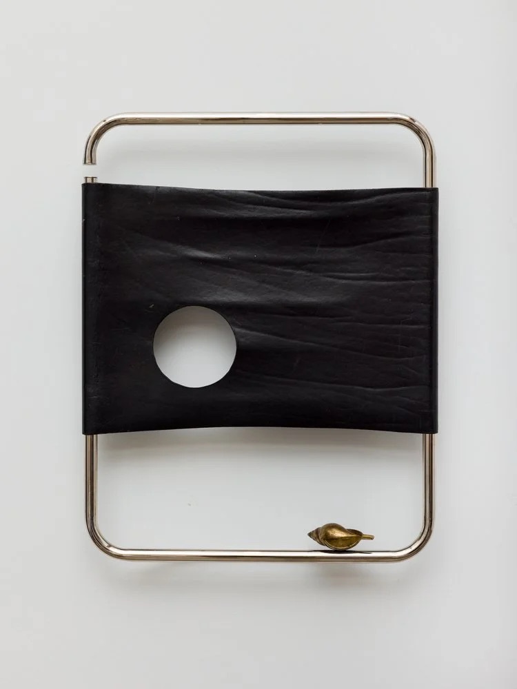 A minimalist object with a rectangular metal frame, a stretched black band featuring a circular hole, and a small brass bird-shaped figure attached to the bottom right corner. The background is white.