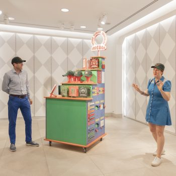 Two people in casual clothing stand near a colorful display cart with hats and Chicago-themed items in a modern, brightly lit room with patterned walls. The woman is speaking while the man listens.