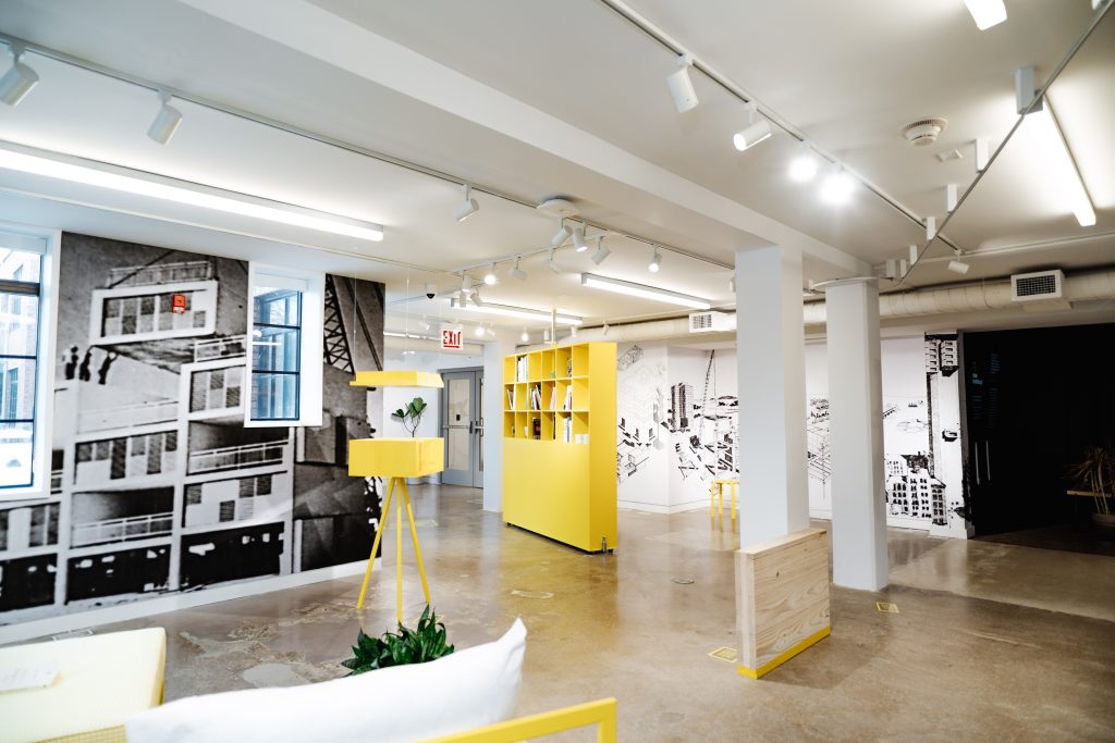 Modern open office space with polished concrete floors, yellow furniture, indoor plants, and large black-and-white cityscape murals on the walls, illuminated by ceiling track lighting.