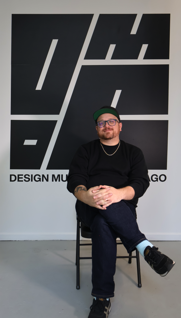 A person with glasses, a beard, and a black cap sits cross-legged on a chair, smiling in front of a large black geometric Design Museum Chicago logo on a white wall.