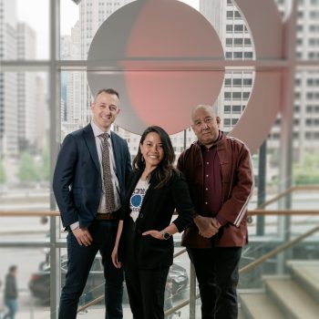 Three people pose together indoors in front of a large circular artwork and tall windows overlooking a cityscape. The group consists of two men and one woman, all dressed in business casual attire.