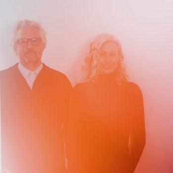 A man and a woman stand side by side, smiling, against a light background. The image is overlaid with a soft red and orange filter, giving it a warm, hazy effect.