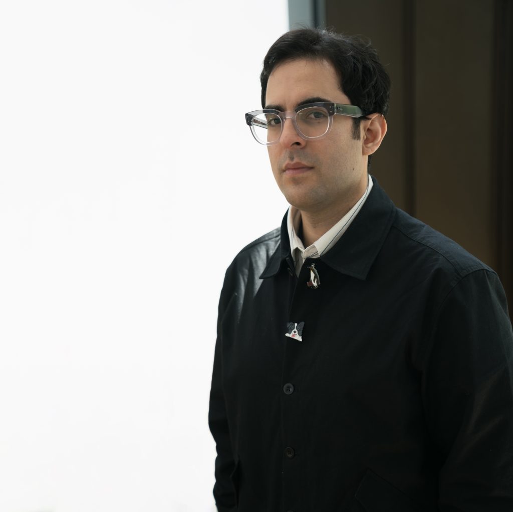 A man with short dark hair and glasses wears a black coat over a white shirt, standing indoors against a bright white background with a dark wall to his right.