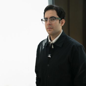 A man with short dark hair and glasses wears a black coat over a white shirt, standing indoors against a bright white background with a dark wall to his right.