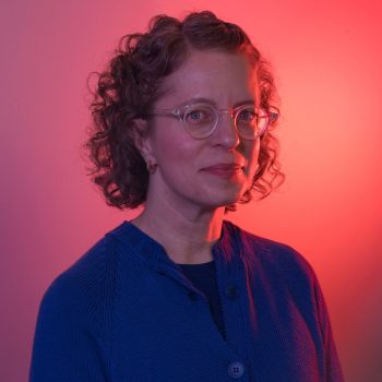 A woman with short curly hair and glasses wears a blue sweater, standing against a pinkish-red gradient background, looking slightly to the side with a neutral expression.