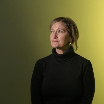 A woman with short brown hair, wearing a black turtleneck, looks thoughtfully to the side. The background fades from green on the left to yellow on the right.