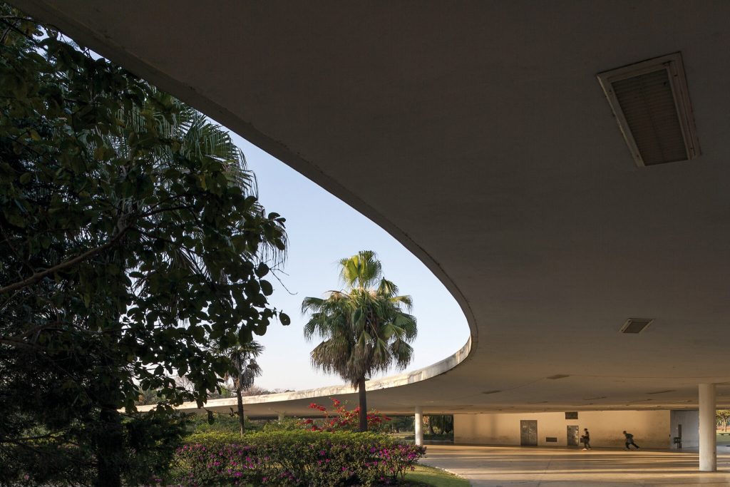 A curved modern building with a large overhang frames palm trees and flowering bushes. Two people walk underneath the overhang, and sunlight illuminates the open space. The sky is clear and blue.
