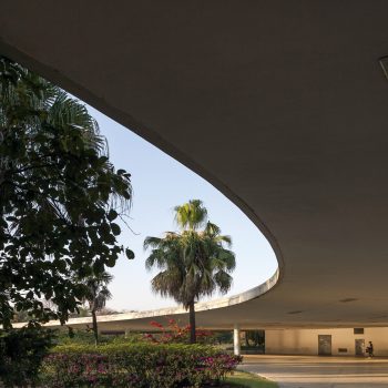A curved modern building with a large overhang frames palm trees and flowering bushes. Two people walk underneath the overhang, and sunlight illuminates the open space. The sky is clear and blue.