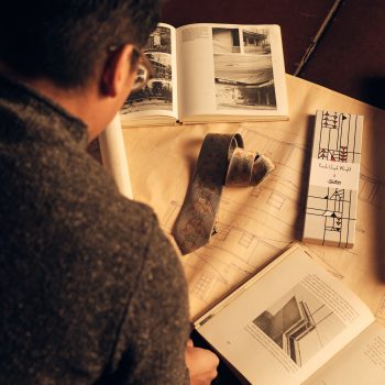 A person views architectural books and drawings spread out on a table, with a rolled blueprint, a patterned tie, and a Frank Lloyd Wright-themed box also visible. Warm lighting creates a cozy atmosphere.