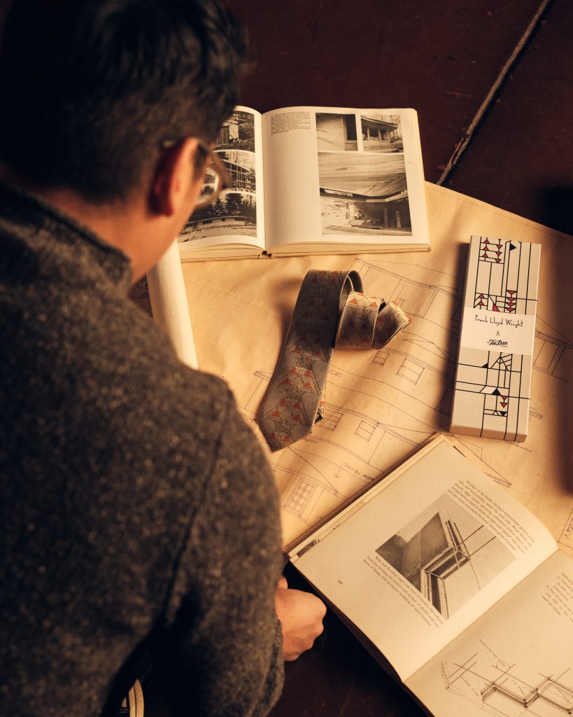 A person views architectural books and drawings spread out on a table, with a rolled blueprint, a patterned tie, and a Frank Lloyd Wright-themed box also visible. Warm lighting creates a cozy atmosphere.