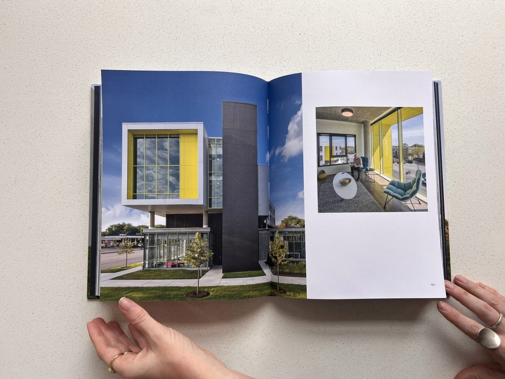 Hands hold open a book displaying modern architecture with bold yellow and black exterior accents on one page, and a person seated on a balcony with yellow framing on the other.