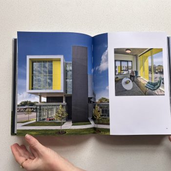 Hands hold open a book displaying modern architecture with bold yellow and black exterior accents on one page, and a person seated on a balcony with yellow framing on the other.