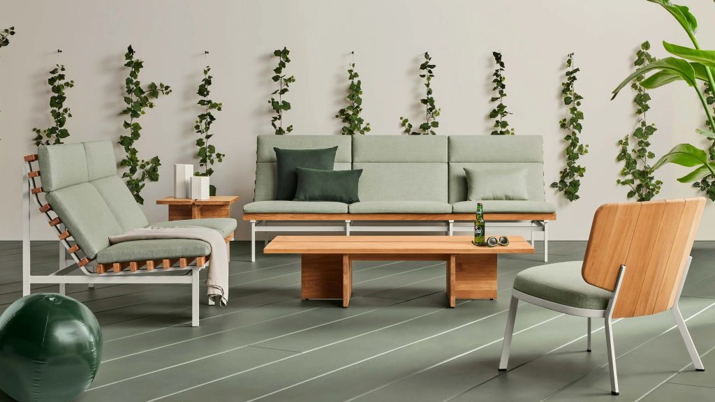 Modern living room with light green cushioned wooden furniture, a wooden coffee table, leafy vines on the wall, and a green exercise ball on the floor. A large plant stands on the right side.