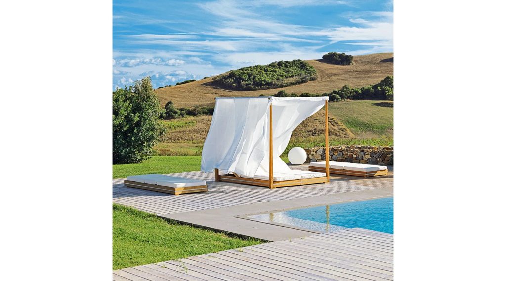 A wooden canopy bed with flowing white curtains sits poolside on a wooden deck, overlooking a grassy landscape and rolling hills under a blue sky with scattered clouds.