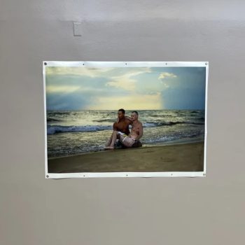 A photo of two men sitting together on a beach at sunset, with waves in the background. The photo is attached to a plain indoor wall.