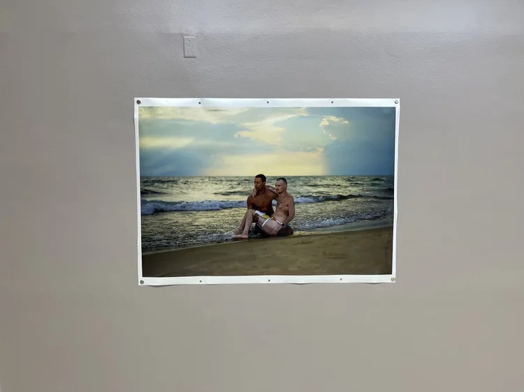 A photo of two men sitting together on a beach at sunset, with waves in the background. The photo is attached to a plain indoor wall.