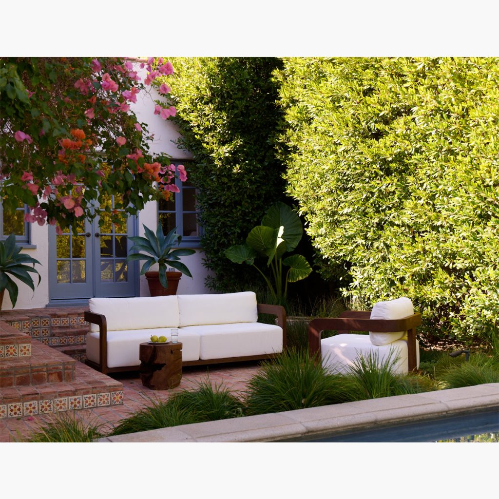 White outdoor sofa and chair with wooden arms are arranged on a tiled patio near a pool, surrounded by green plants, trees, and colorful flowers. Blue French doors lead to the house in the background.