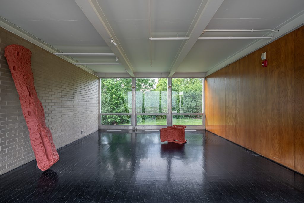 A modern, minimalist room with large windows, dark shiny floor, beige brick and wood-paneled walls, and two abstract red sculptures—one leaning against the wall, the other placed in the center. Green trees are visible outside.