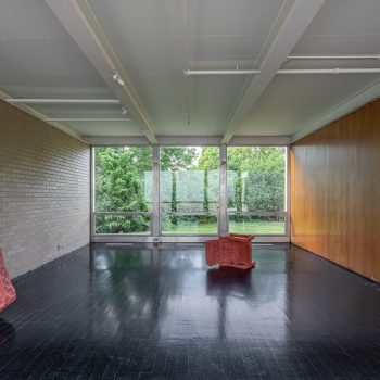 A modern, minimalist room with large windows, dark shiny floor, beige brick and wood-paneled walls, and two abstract red sculptures—one leaning against the wall, the other placed in the center. Green trees are visible outside.