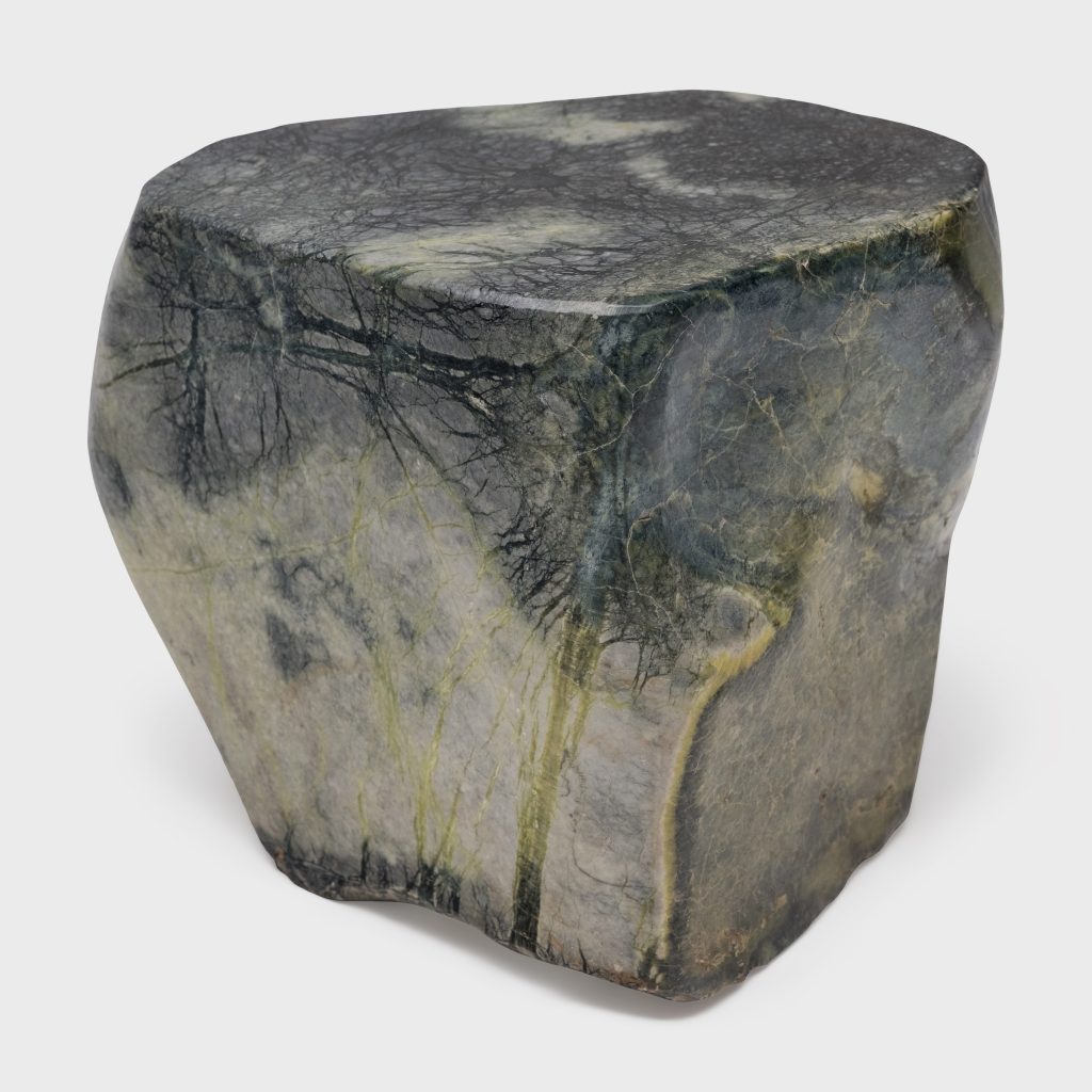 A large, irregularly shaped stone with a smooth surface, dark gray and green hues, and visible natural veining patterns, sits on a plain white background.