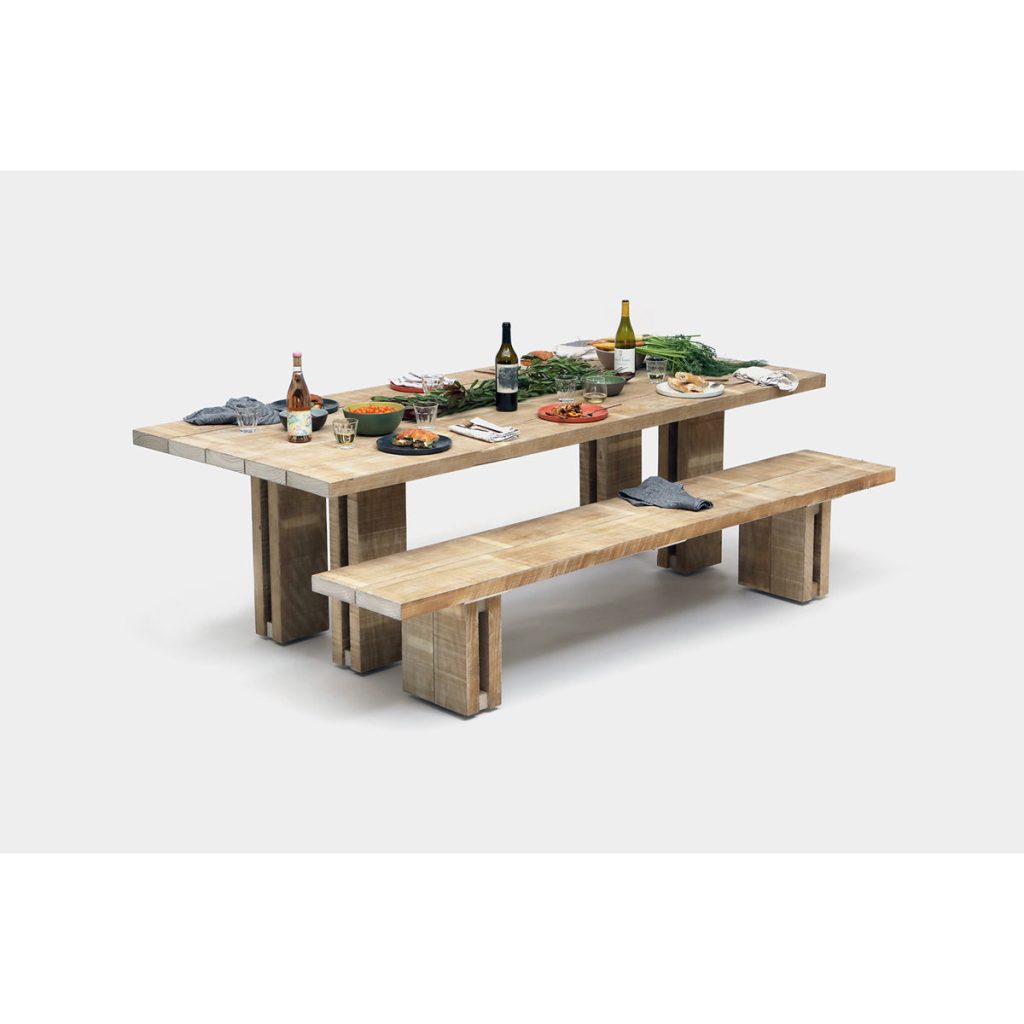 A wooden dining table with matching bench displays food, wine bottles, plates, and utensils, set against a plain white background.