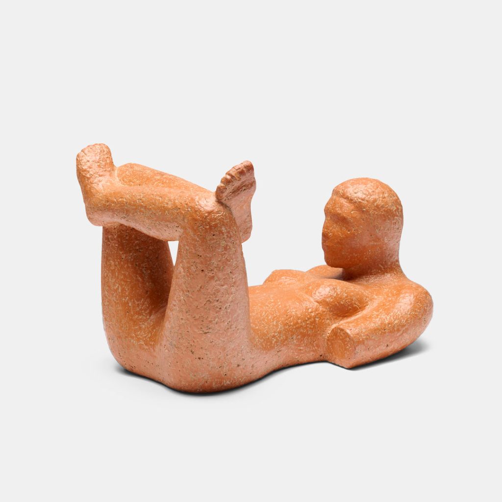 A textured, orange-brown sculpture of a reclining, nude human figure resting on its back with bent legs raised and arms by its sides, set against a plain, light background.