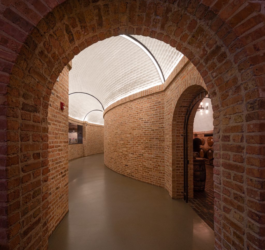 Curved brick hallway with arched ceilings and smooth flooring, leading to a room with barrels. Warm lighting highlights the textured brick walls and the white, vaulted ceiling above.