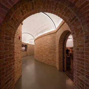 Curved brick hallway with arched ceilings and smooth flooring, leading to a room with barrels. Warm lighting highlights the textured brick walls and the white, vaulted ceiling above.